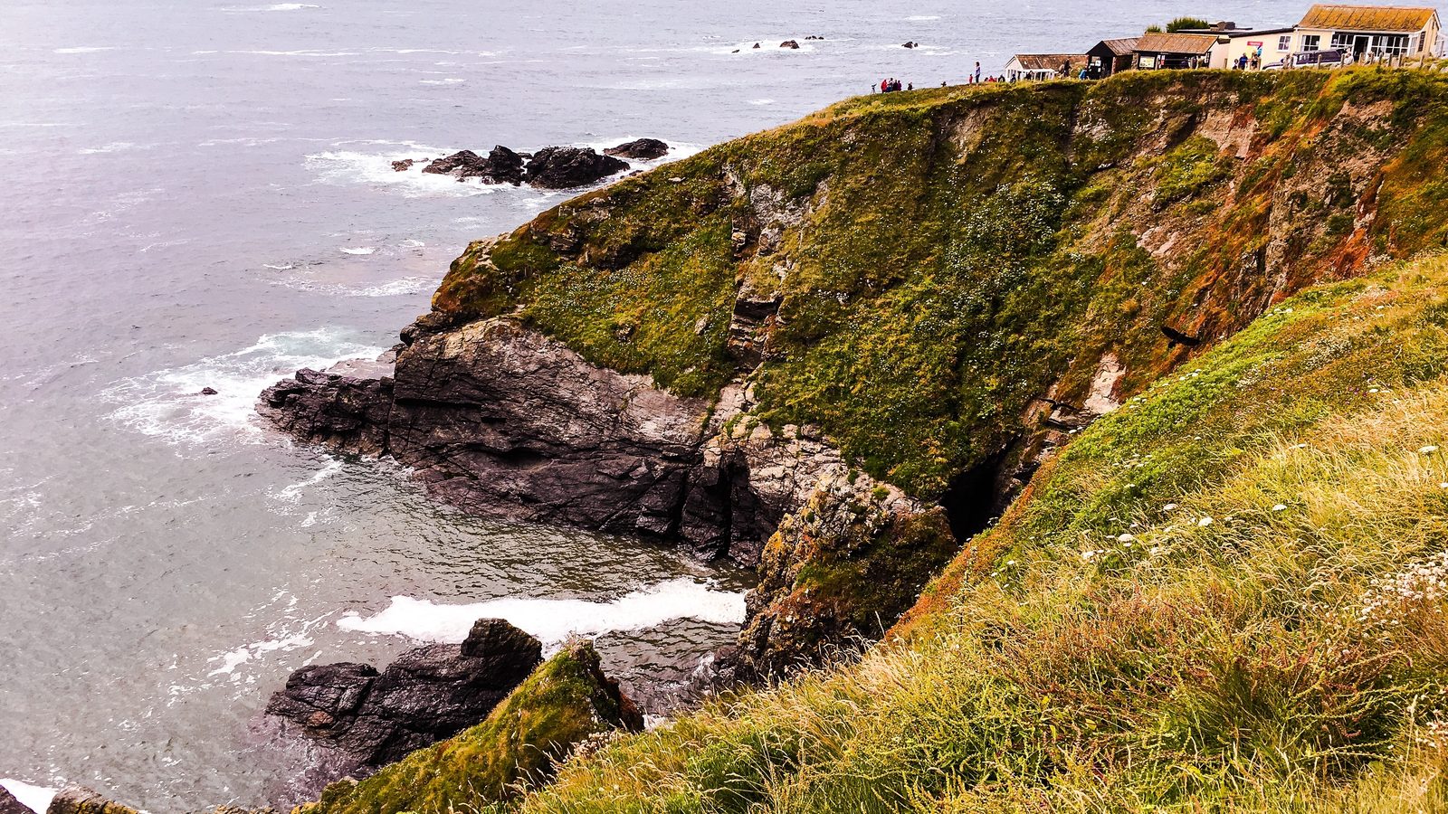 Grassy cliff-top path above dark serpentine rocks and surf at Lizard Point, with buildings on the headland