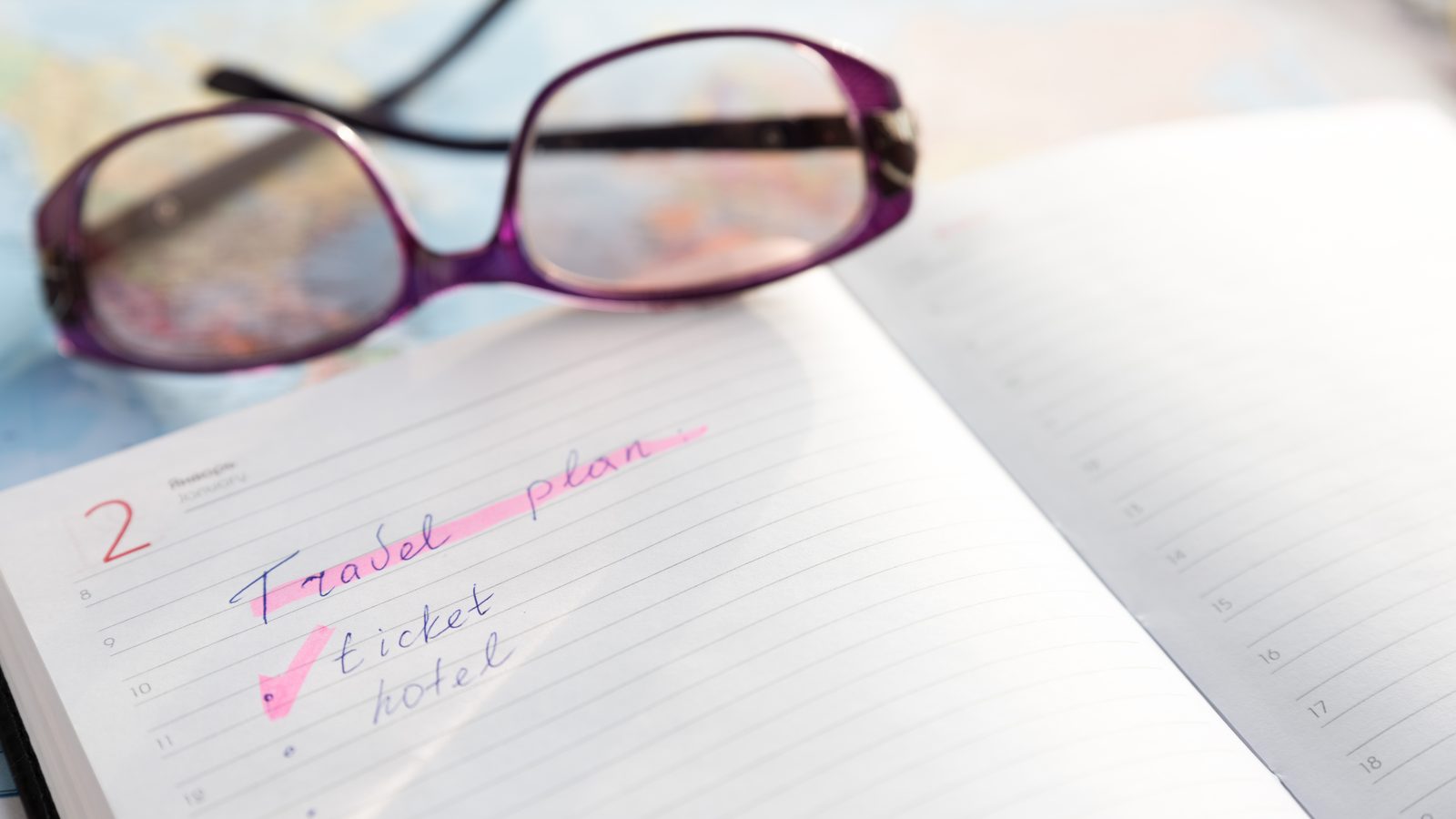 A handwritten travel-plan checklist in an open diary, with reading glasses resting on a blurred map behind it