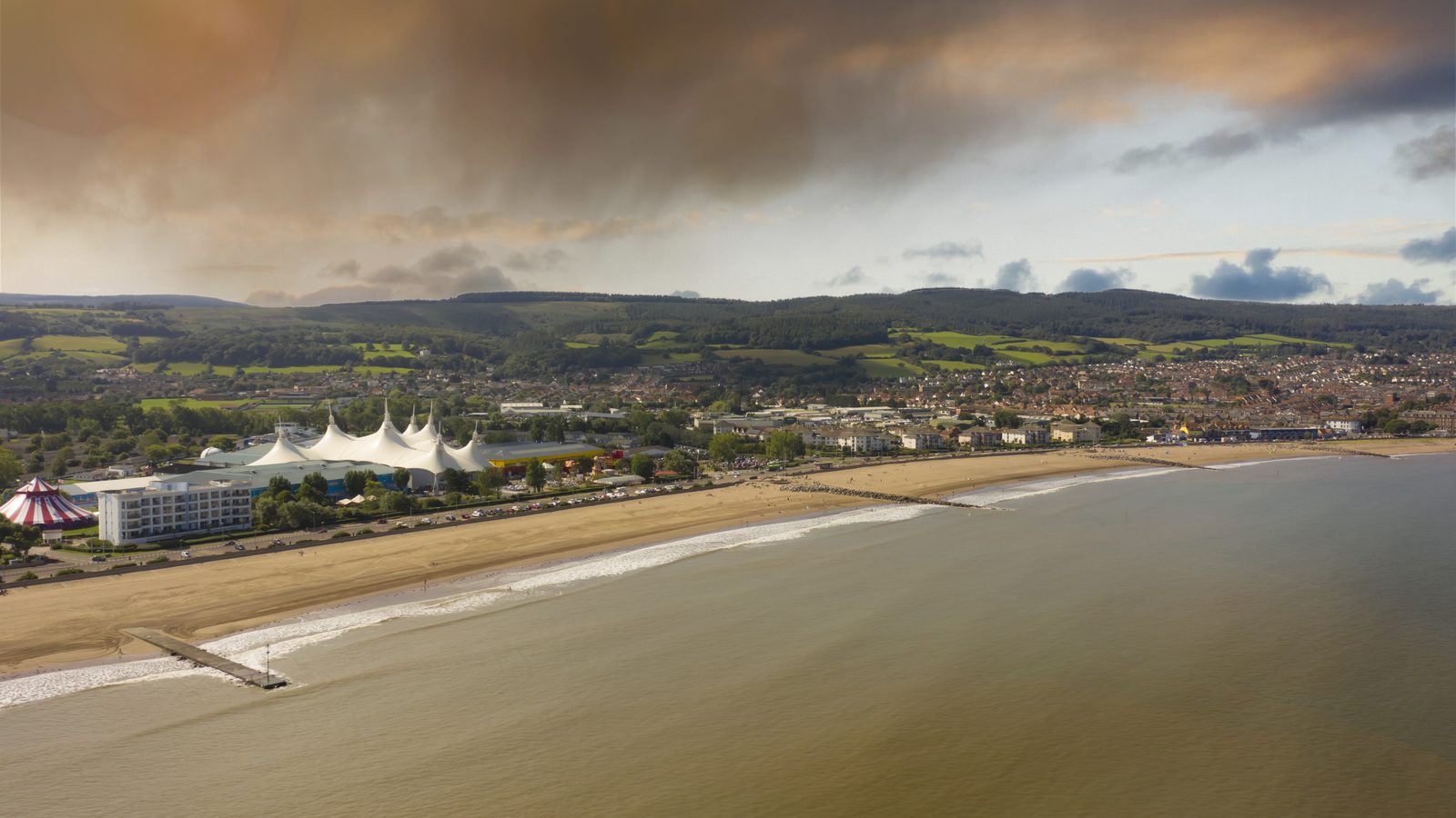 Aerial view of Minehead seafront under a cloudy sky, with the beach, the white tented roofs of the holiday park and the wooded Exmoor hills behind the town