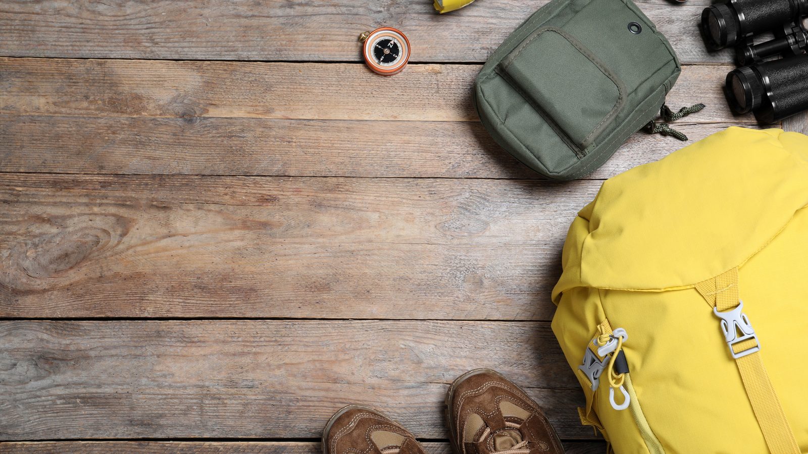 Flat lay on weathered boards of a yellow rucksack, walking boots, a compass, a torch, a pocket knife, binoculars and a small green pouch