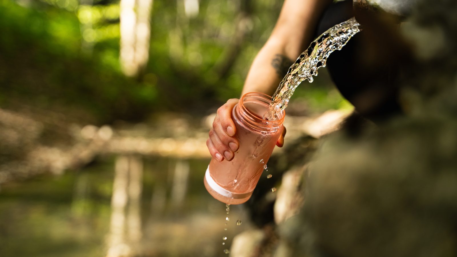 A walker filling a pink water bottle from a stone spring spout in dappled woodland, with a stream out of focus behind